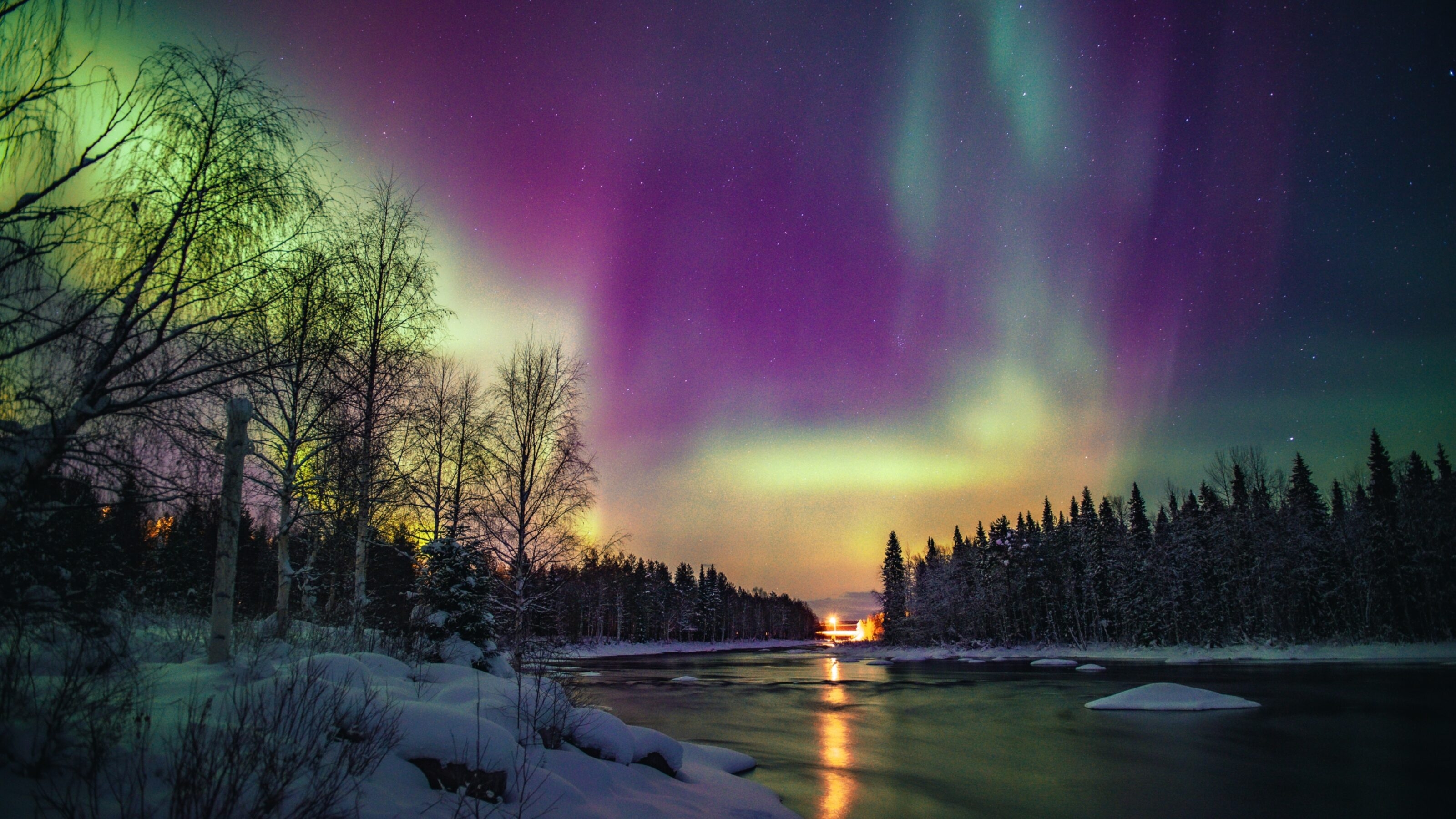 Aurora borealis over a snowy forest with a river reflecting colorful lights in the night.
