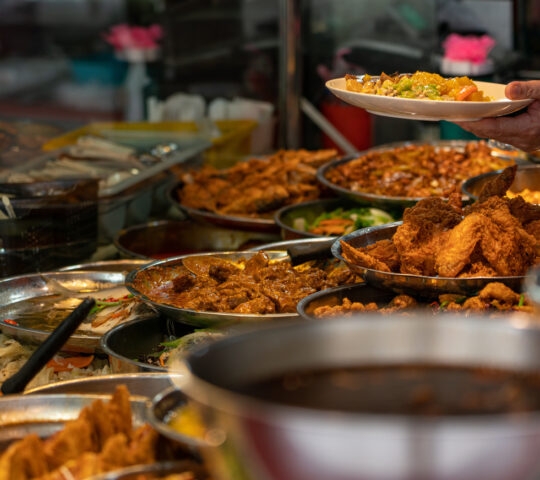 Person serving food at a buffet with various dishes in the foreground.