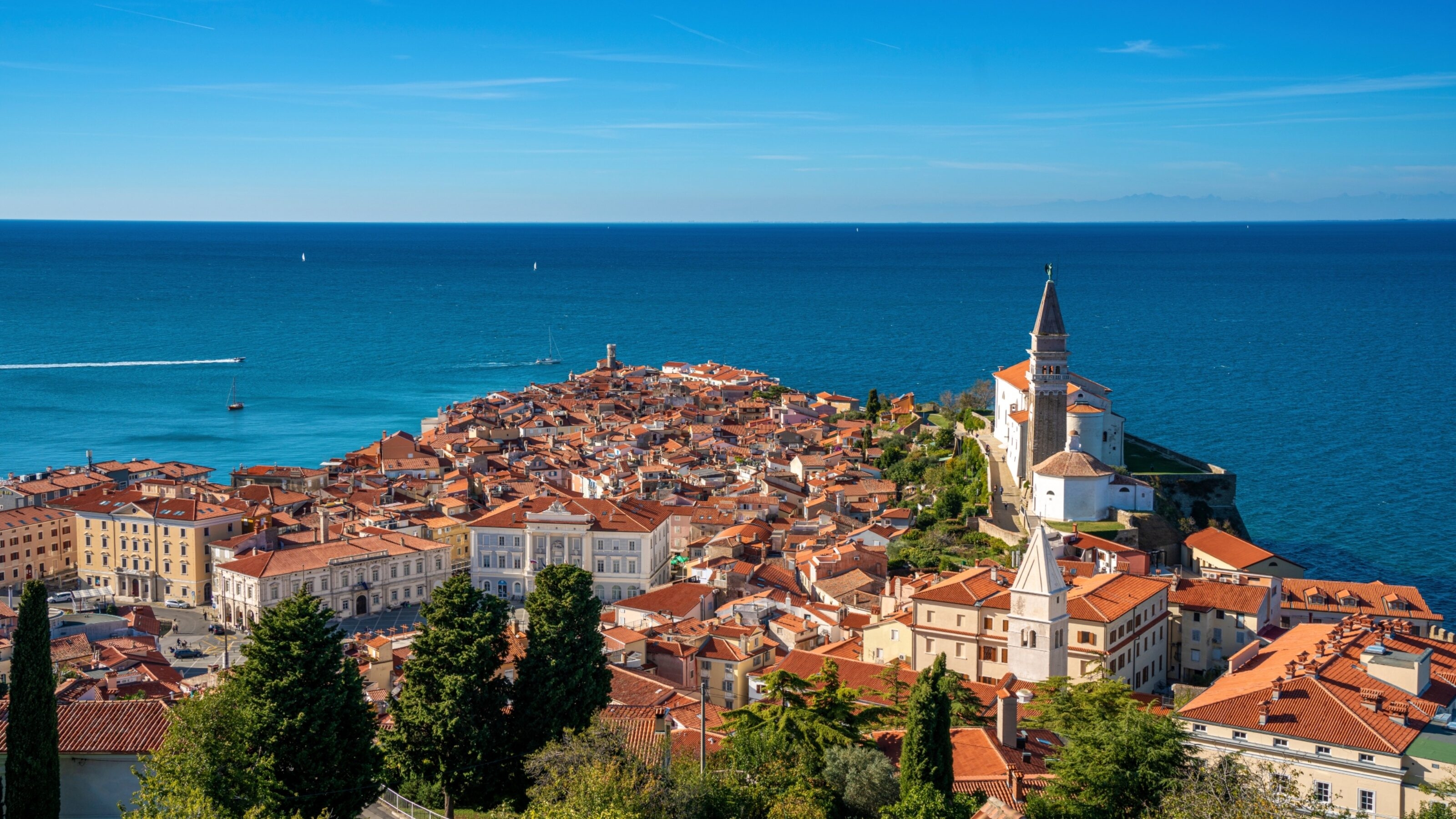 A coastal town with terracotta rooftops, a prominent church spire, and a clear blue sea in the background.