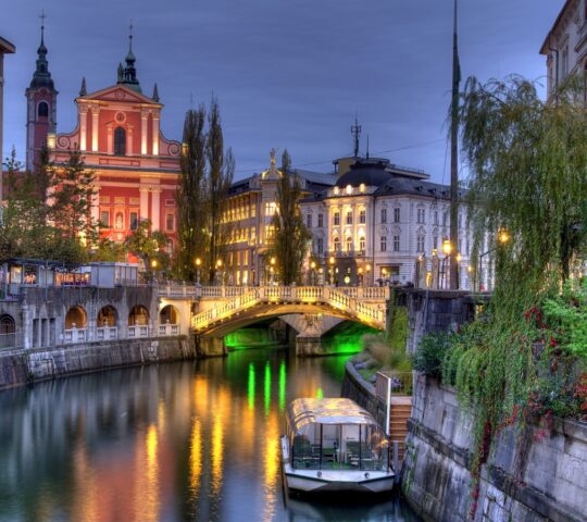 Twilight cityscape with illuminated buildings, a bridge, and a boat on the river.