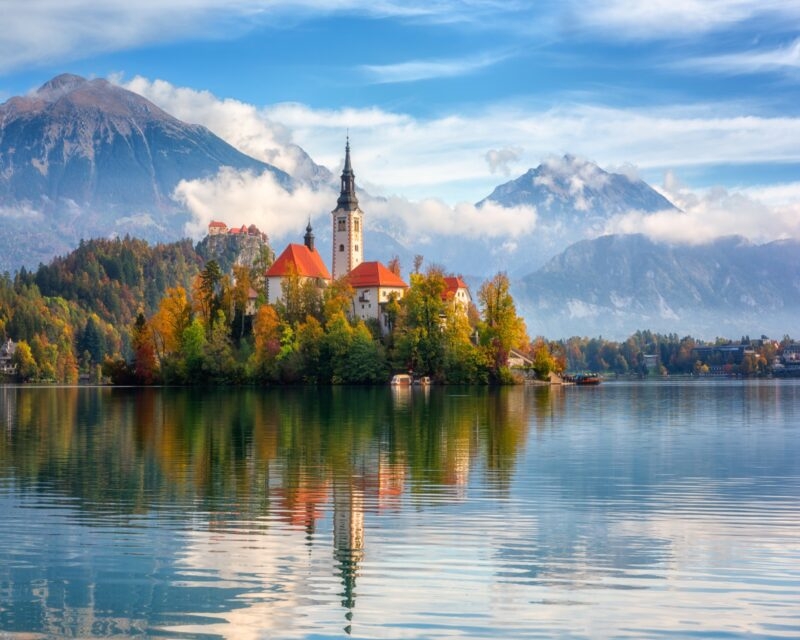 Island church with red roof in a clear lake with mountains and clouds in the background.