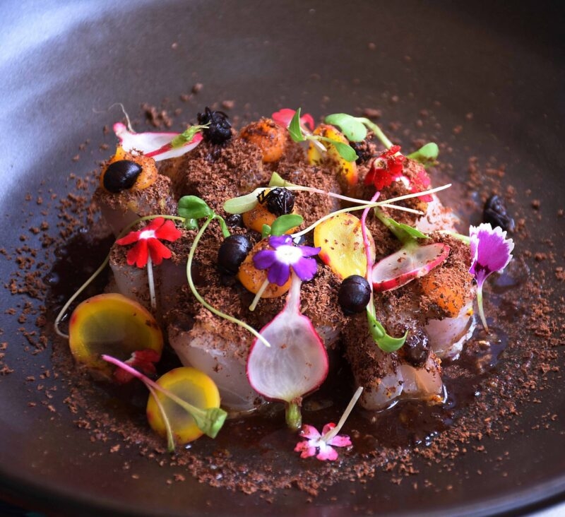 Gourmet dish with edible flowers, herbs, and chocolate soil on a dark plate.