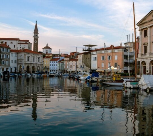 Sunset over a tranquil harbor with boats and old European buildings reflecting in the water.