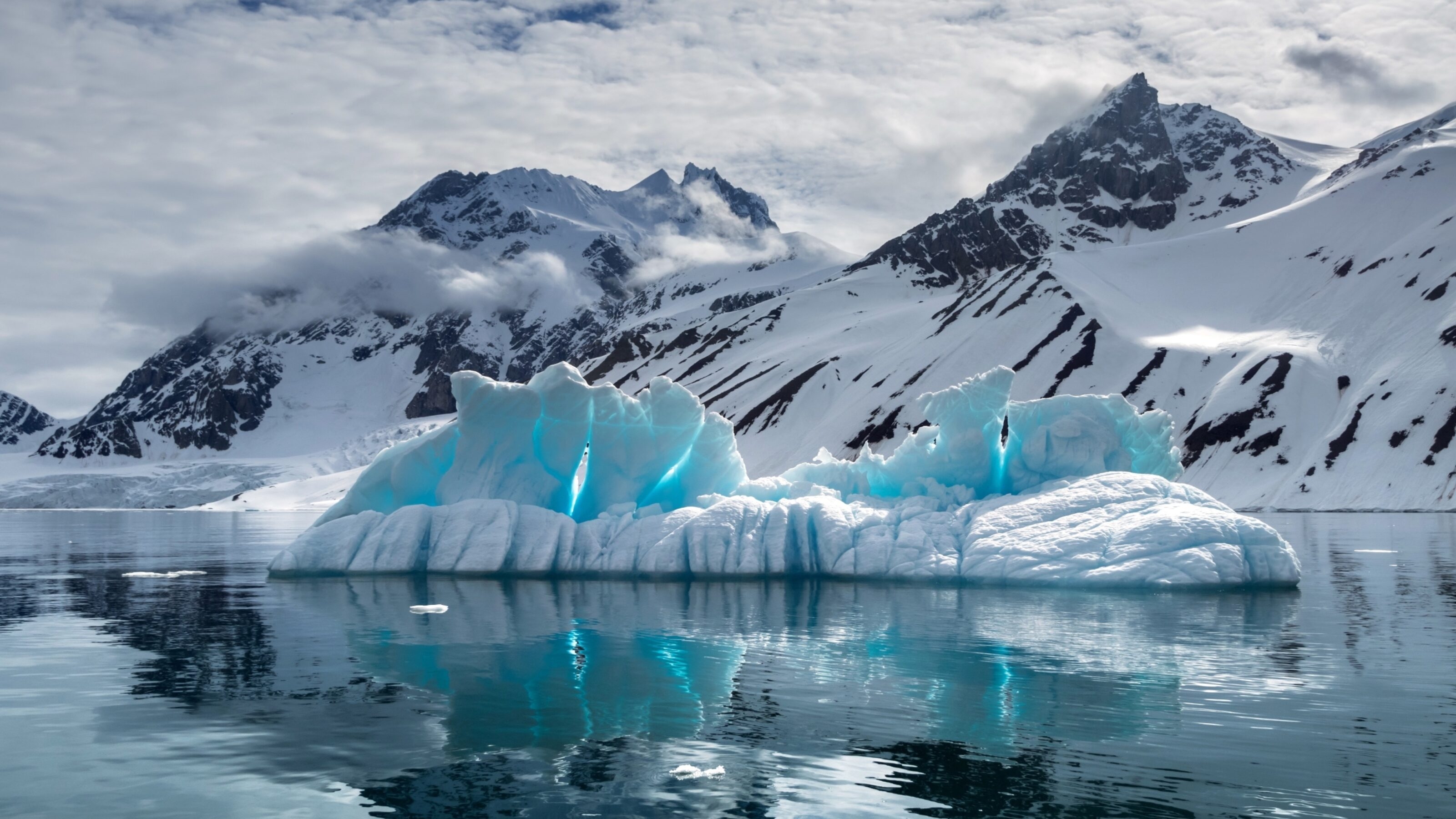 A vivid blue iceberg floats before snow-capped mountains under a cloudy sky.