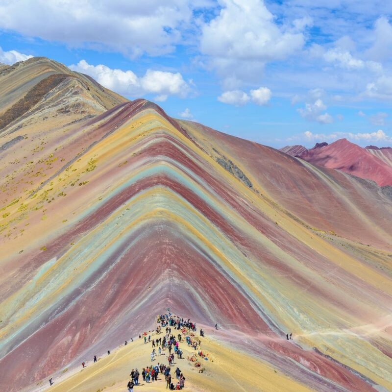 Colorful striped mountains with a crowd of visitors at the base under a blue sky.