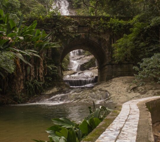 Stone arch bridge over a stream leading to a waterfall surrounded by lush greenery.