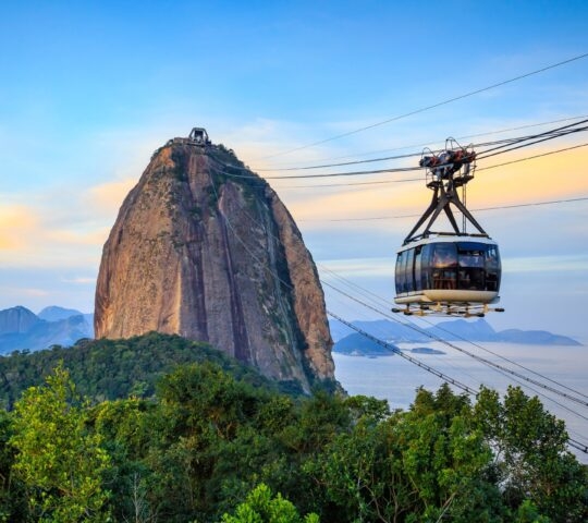Aerial view of a lush coastal village with boats in a blue bay flanked by green hills.
