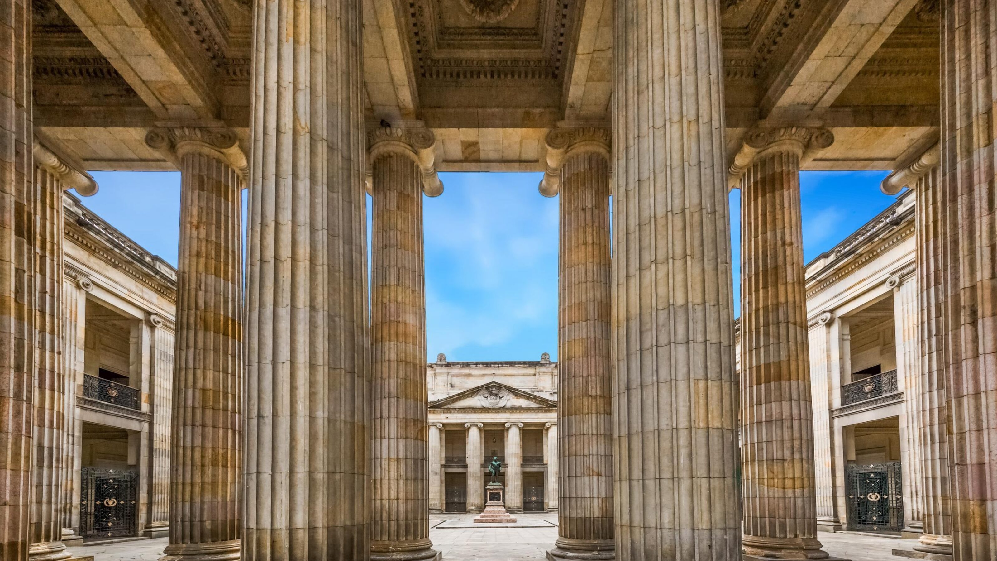 View through tall neoclassical columns to a courtyard with a statue and blue sky above.