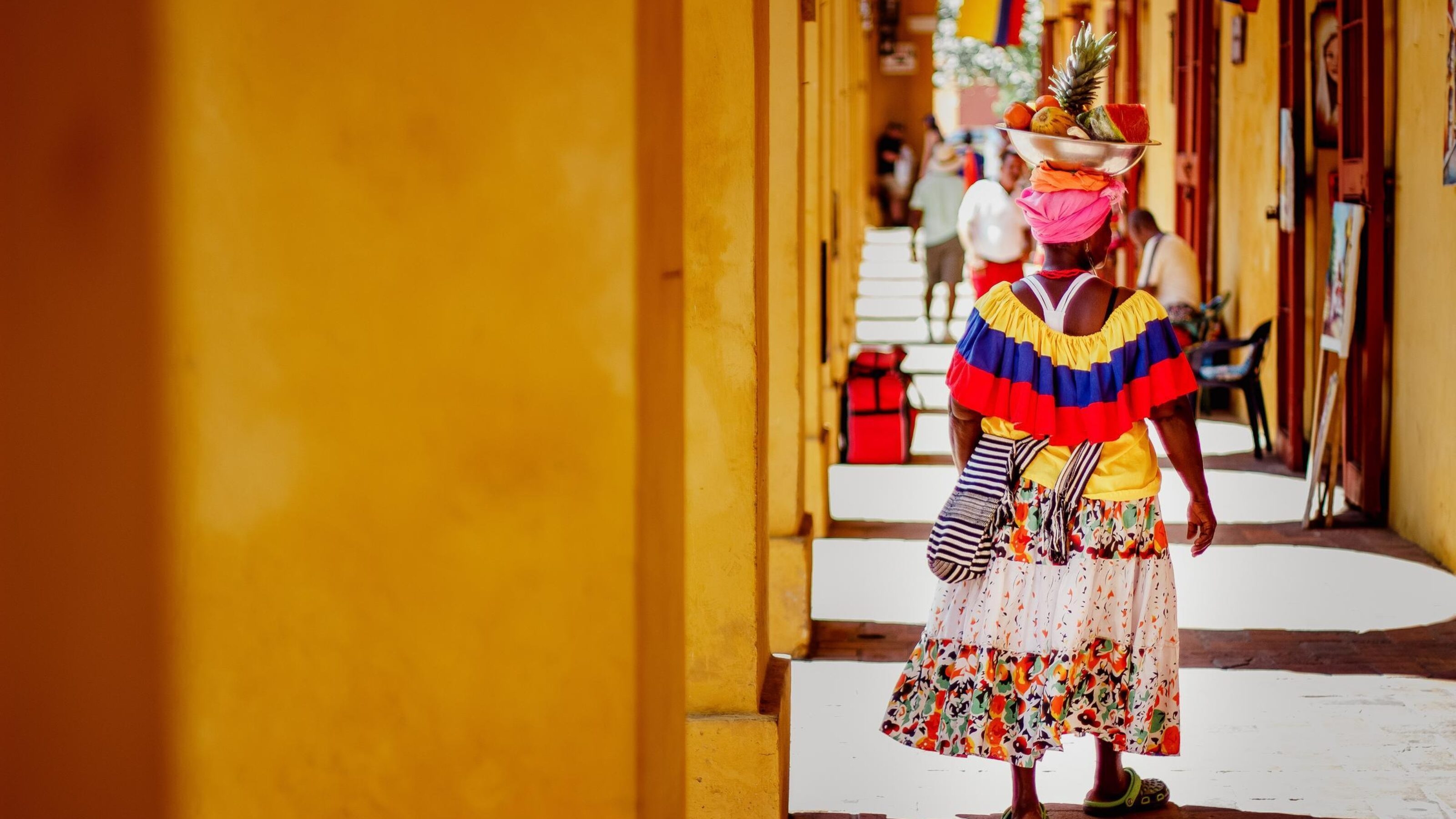 Person in colorful traditional attire carrying fruit on head down a sunny, colonial arcade.