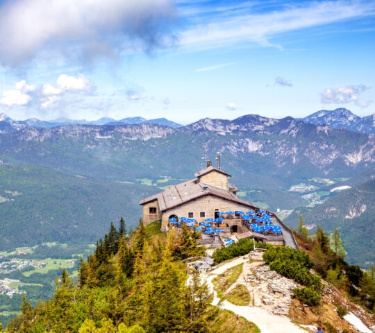 Mountain lodge with blue umbrellas overlooking alpine scenery.