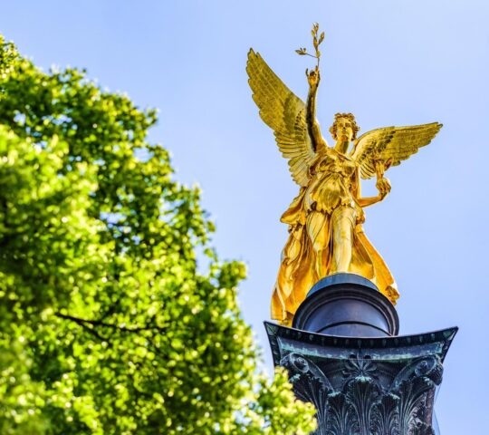 Golden angel statue against blue sky, surrounded by green foliage.