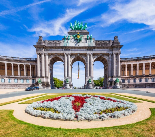 A grand triumphal arch with sculptural details and a colourful floral foreground under a blue sky.