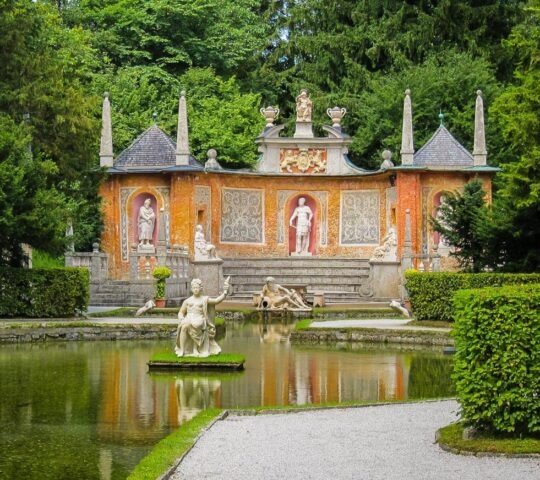 Ornate garden with sculptures, a pond, and a decorative wall amidst green foliage.