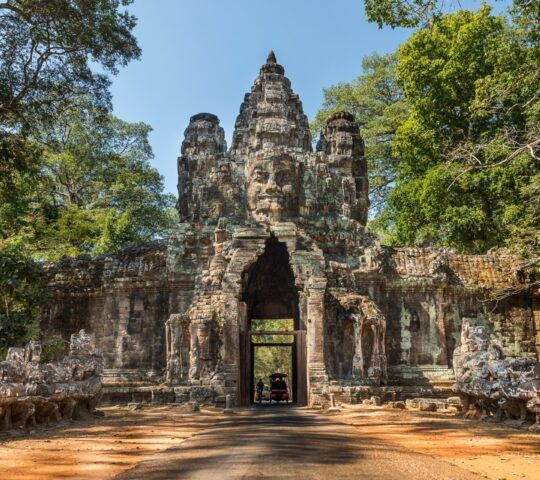Ancient stone gate with a large carved face at Angkor Thom, surrounded by trees under a blue sky.