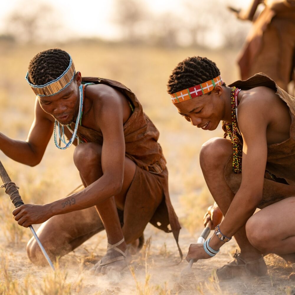Two individuals in traditional attire crouching with spears on a grassy savannah.