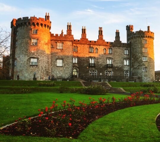 A historic stone castle with turrets bathed in golden sunlight, surrounded by vibrant green lawns and red flowers.