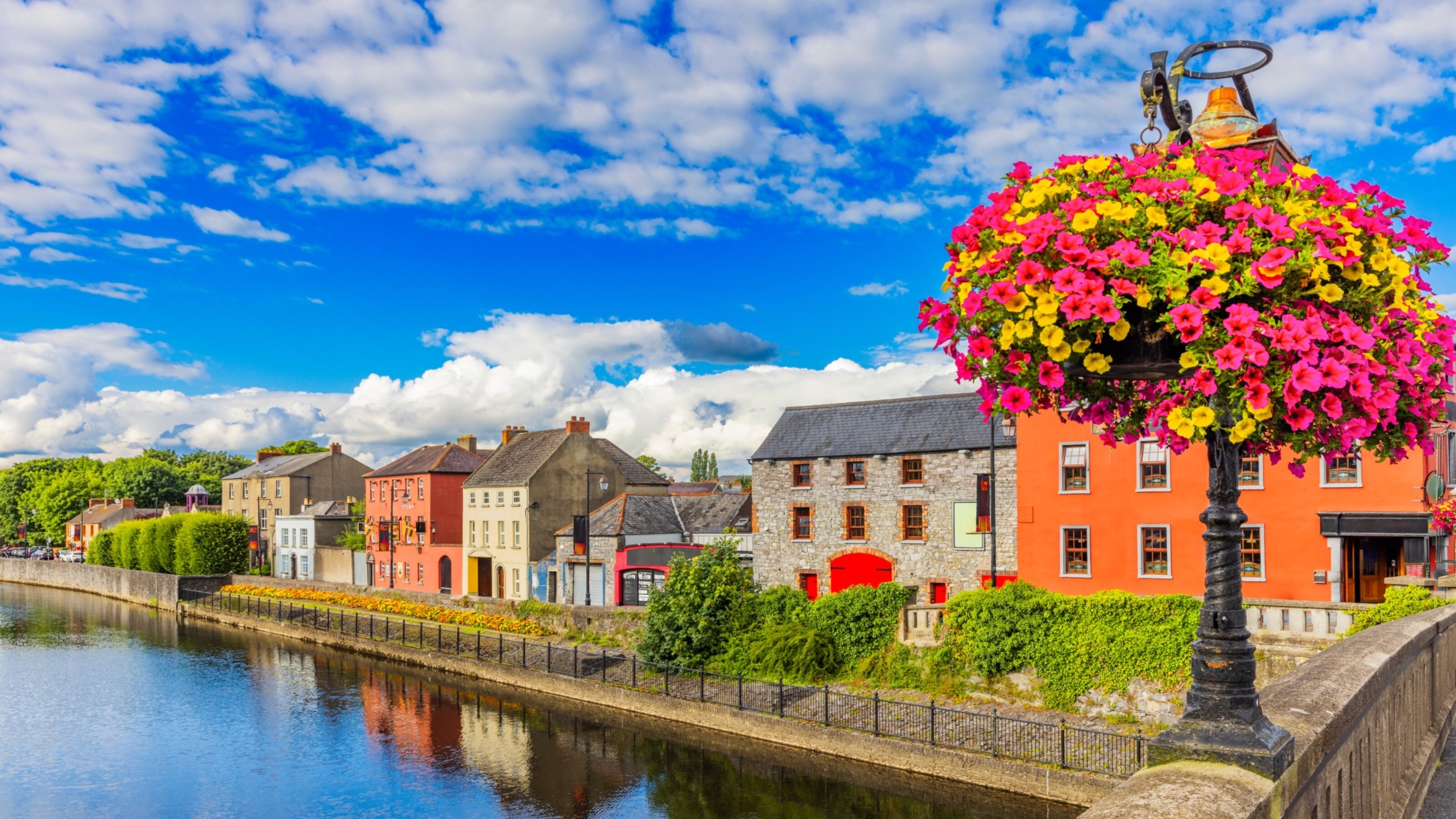 Colourful flowers atop a lamp post with a scenic view of a river and vibrant buildings under a blue sky.