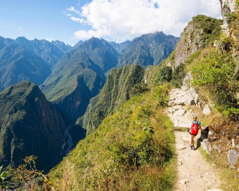 A person in a red backpack hikes on a narrow mountain path overlooking a deep, lush green valley and towering mountains, a highlight of luxury Machu Picchu holidays.