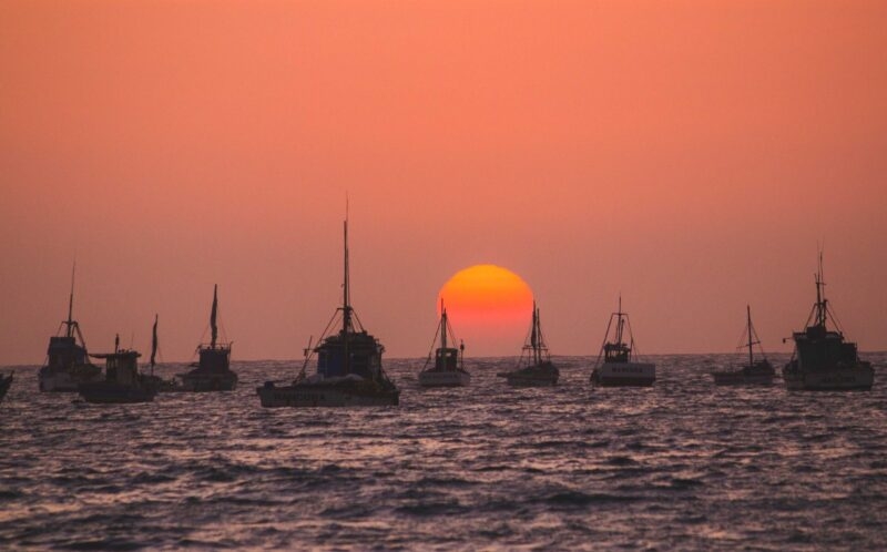 Sunset over the ocean, silhouetting fishing boats against a vivid orange sky.