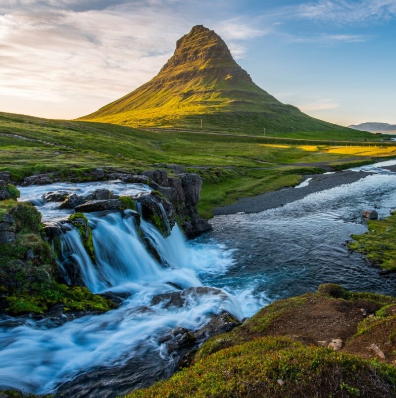 Waterfall with a flowing river in the foreground and a distinct mountain in the background at sunset.