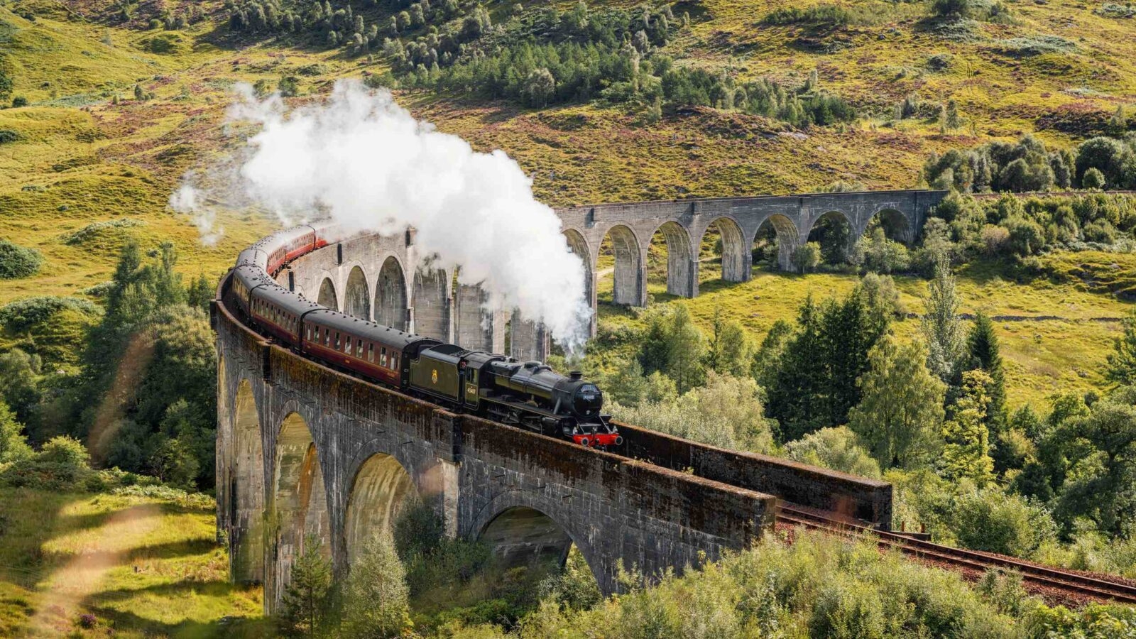 Steam train on Glenfinnan Viaduct amid green hills.