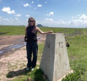 Person standing next to a Kenya-Tanzania border marker in a grassy savanna.