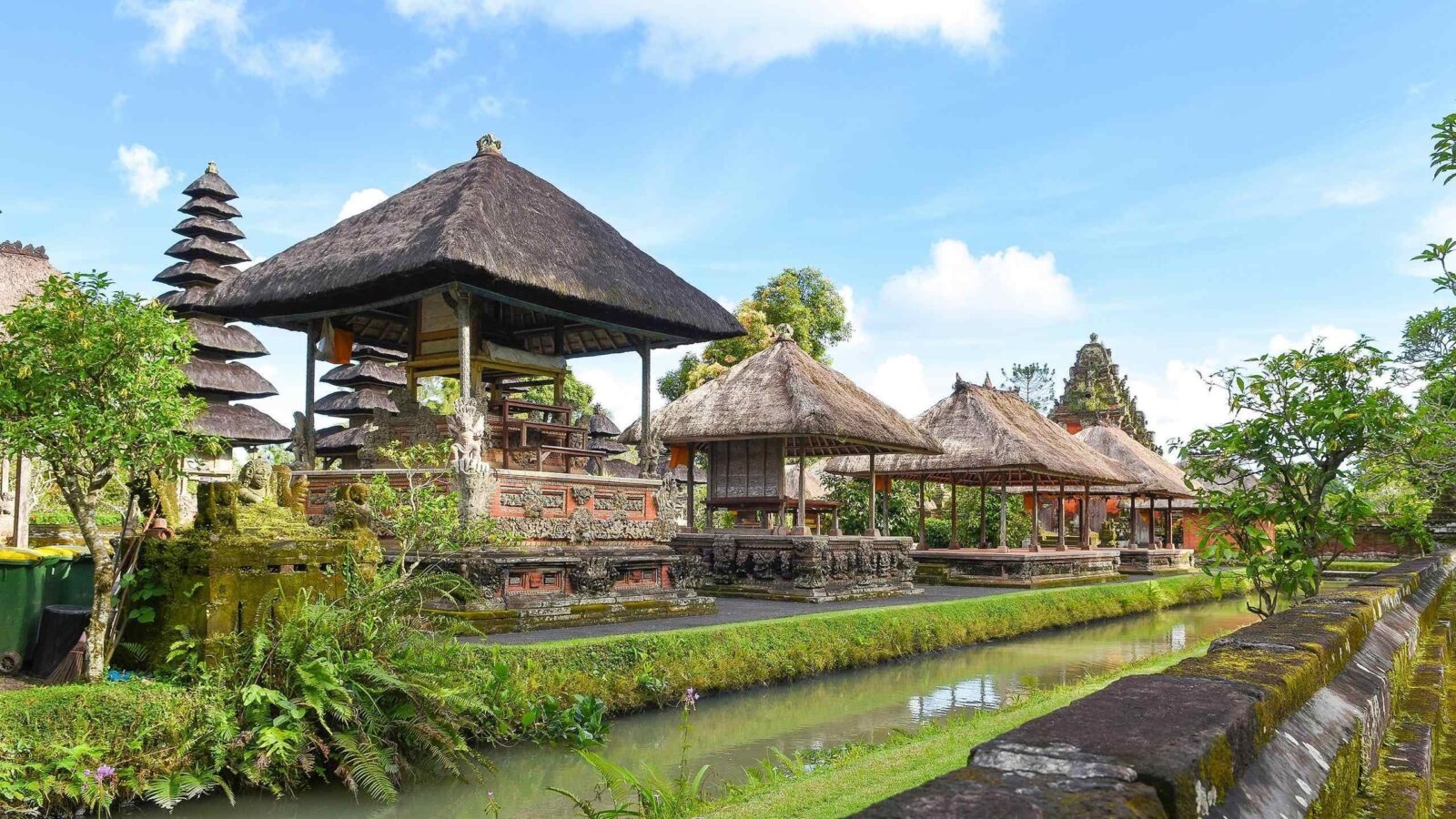 Traditional Balinese architecture featuring thatched-roof structures, intricate stone carvings, and lush greenery beside a tranquil waterway.