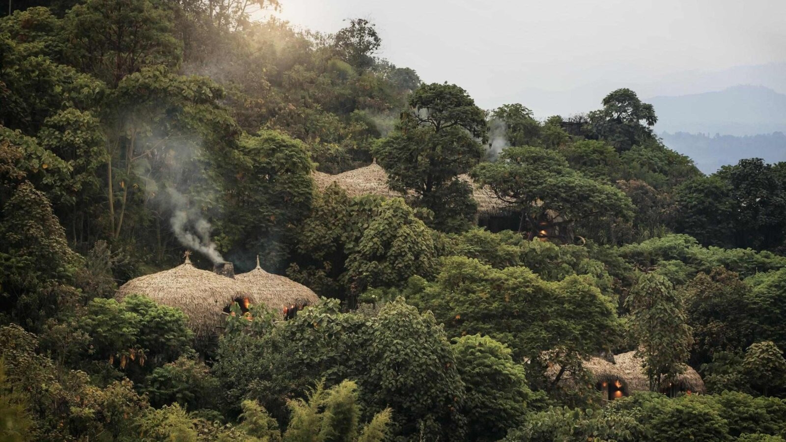 A hillside village of thatched huts hidden among lush greenery, with smoke gently rising from chimneys, illuminated by soft sunlight.