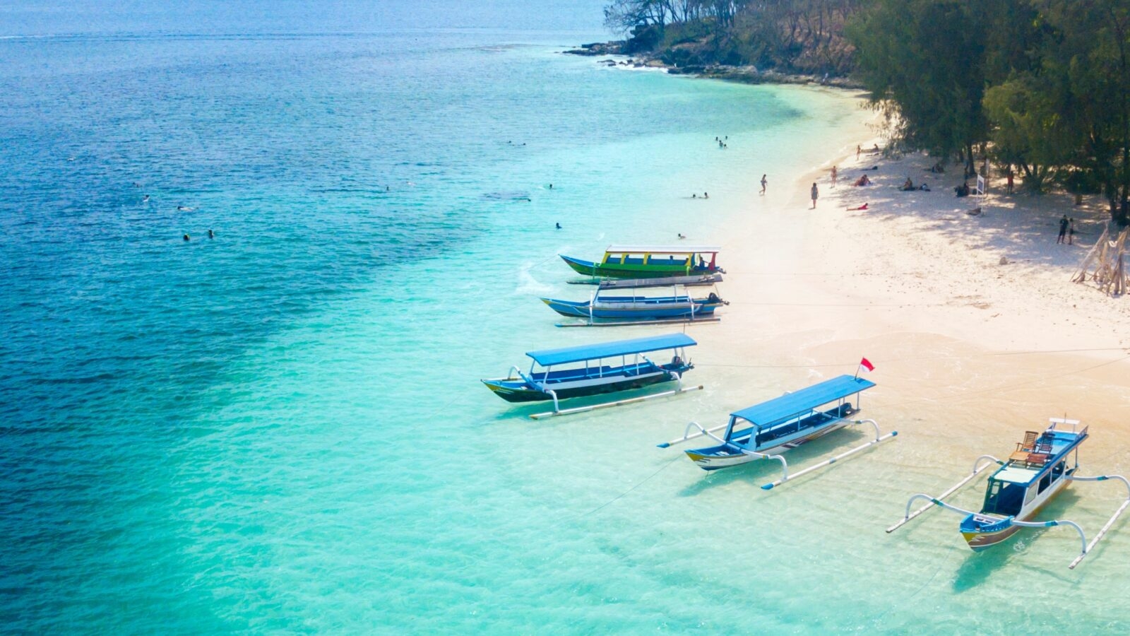 Colorful boats anchored on a tranquil beach with clear turquoise water and people enjoying the sun and water activities.