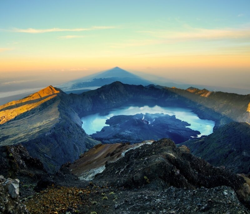 A stunning view of a volcanic crater at sunrise, featuring a tranquil blue lake surrounded by rugged mountains and soft shadows.