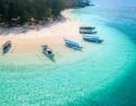 Aerial view of a serene beach with turquoise waters and several traditional boats anchored near the shore, surrounded by green trees.