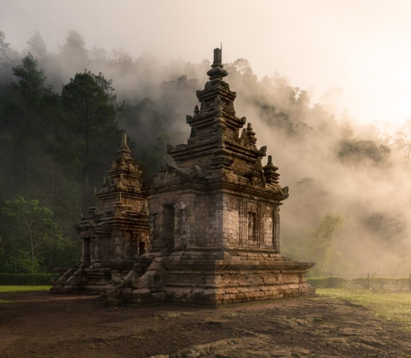 Ancient stone temple shrouded in mist, surrounded by lush greenery and silhouetted against a hazy morning sky.