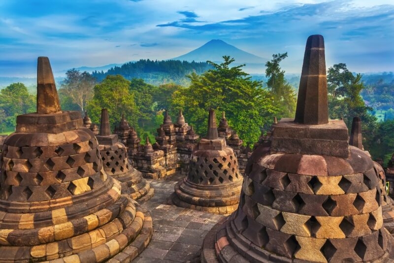 Ancient stone stupas at Borobudur Temple, surrounded by lush greenery and a distant volcanic mountain under a blue sky.