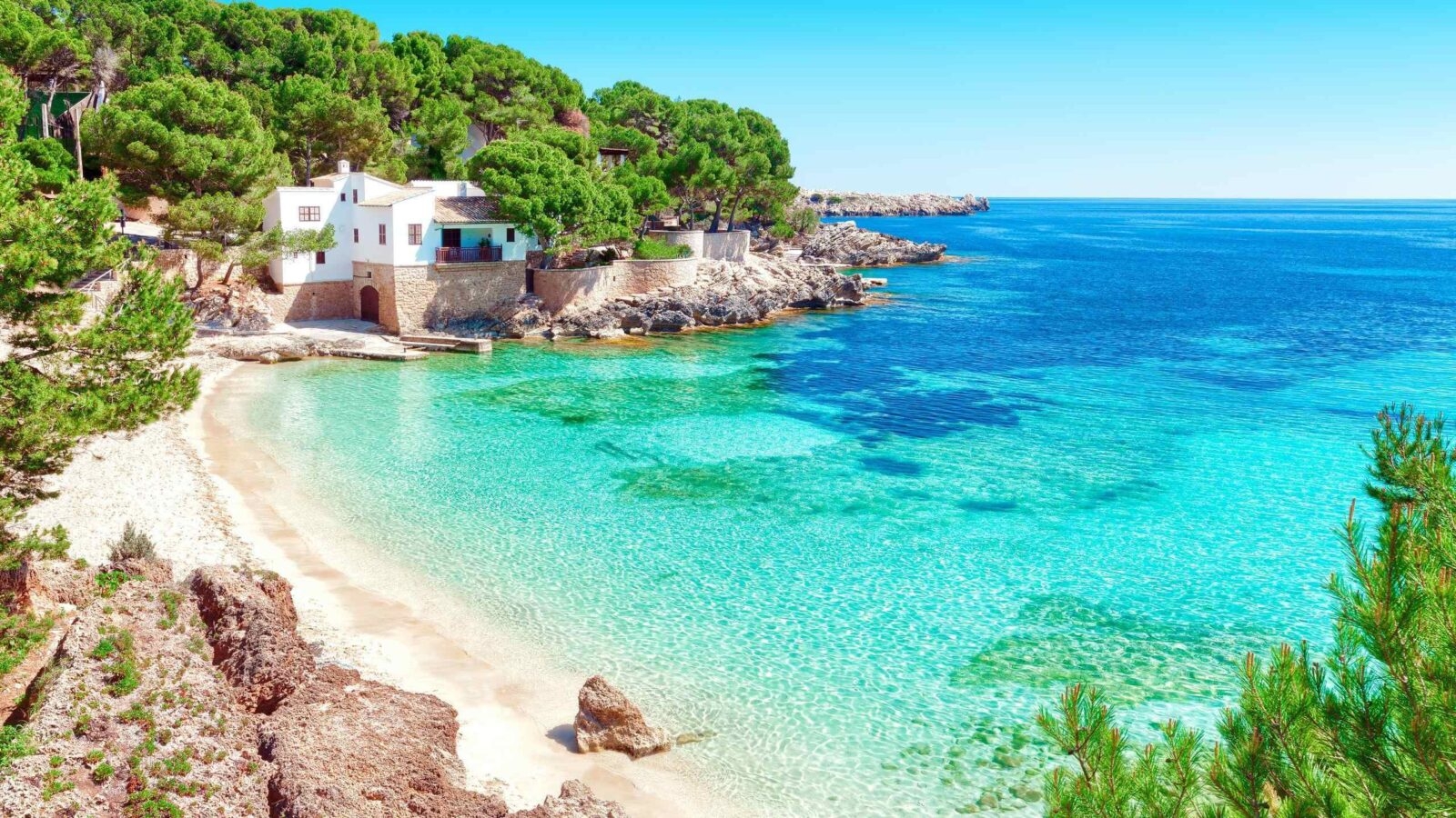 A tranquil coastal scene featuring a white house nestled among lush greenery, with crystal-clear turquoise waters and a sandy beach.