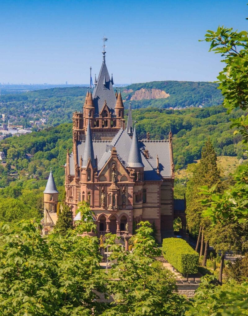 A stunning, ornate castle surrounded by lush greenery, with pointed roofs and decorative towers, under a clear blue sky.
