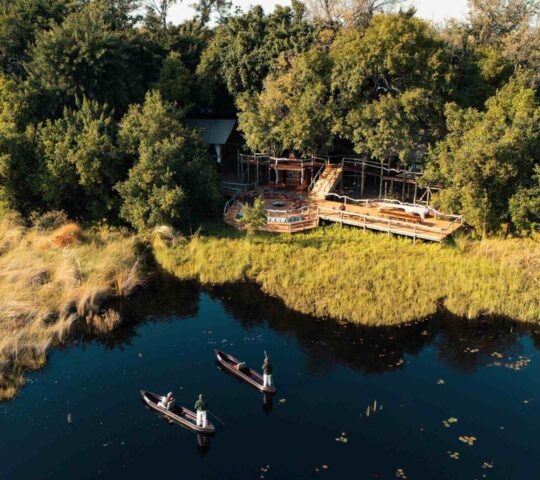 Aerial of Shinde Camp which sits at the edge of a lagoon, where two men steer a pair of traditional canoes.
