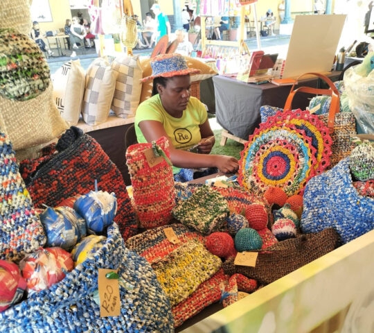 A vendor sits behind a table filled with brightly colored, patterned woven bags and round textile crafts at a market.
