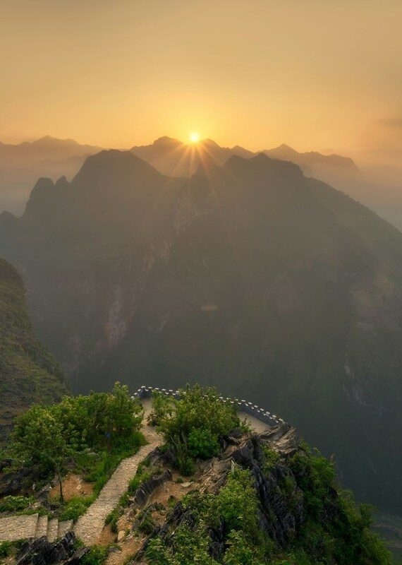 A sunset view over a vast landscape of misty, layered mountains from a high green overlook with a path, a stunning sight for luxury Machu Picchu vacations.