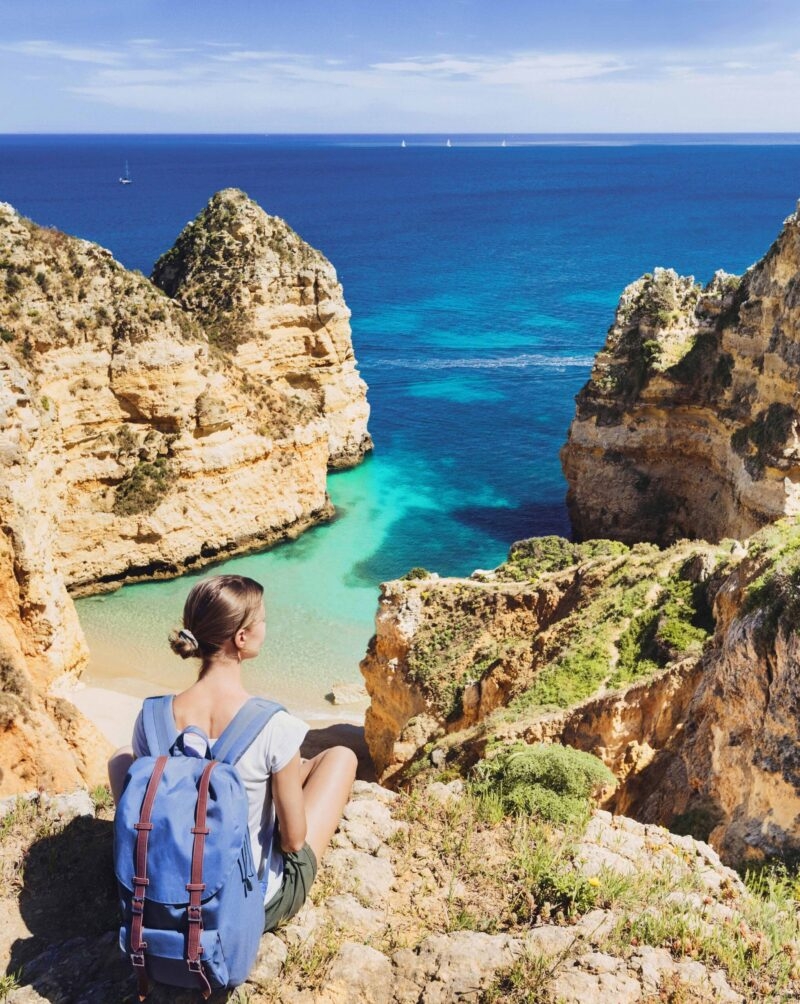 Woman sitting on a cliff overlooking a hidden cove with turquoise water and rock formations, a view from luxury Portugal tours.