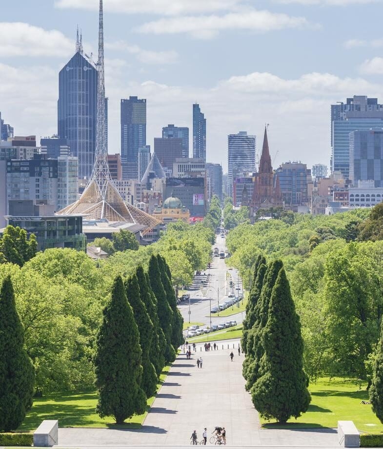 The tree-lined boulevard leading into Melbourne’s skyline and Perth’s city skyline reflected on the Swan River under soft evening light.