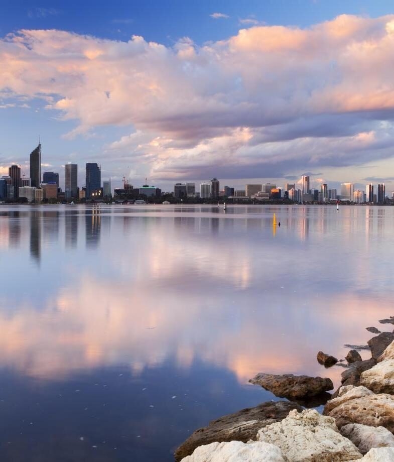 The tree-lined boulevard leading into Melbourne’s skyline and Perth’s city skyline reflected on the Swan River under soft evening light.