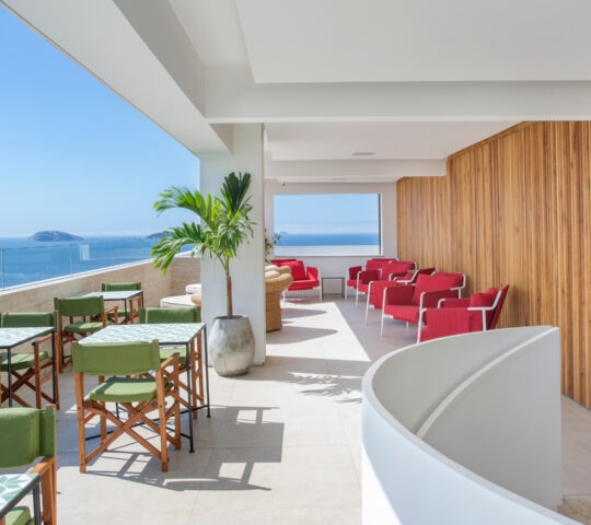 An open-air terrace with white ceiling and floor. On the right are red lounge chairs, on the left are green chairs and dining tables. In the background is a clear glass railing and beyond is the blue sea and sky with a few islands in the distance.
