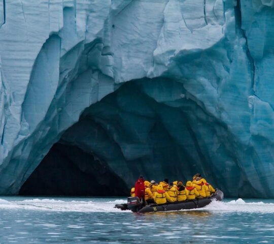 A group of people on a Zodiac excursion in the Arctic, cruising past a cave carved into a giant glacier.