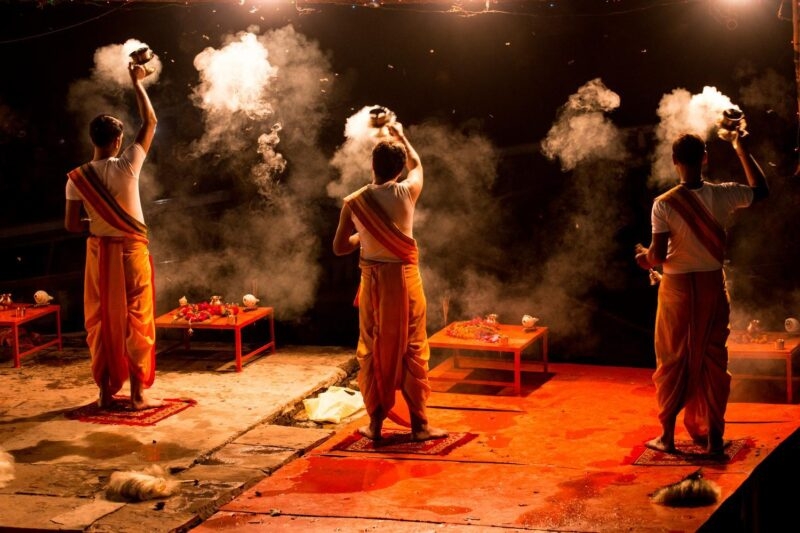 Three monks dressed in orange holding flaming lanterns in the darkness as part of an Aarti ceremony in Varanasi