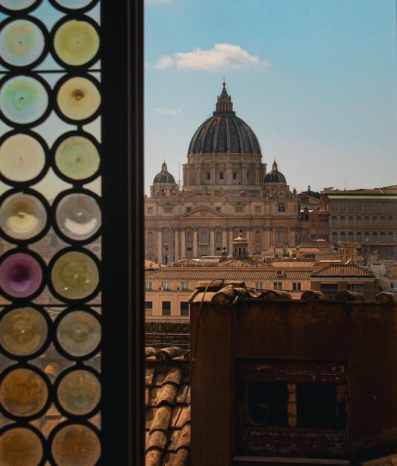 View through an open stained glass window over rooftops and St Peter's Basilica in Rome