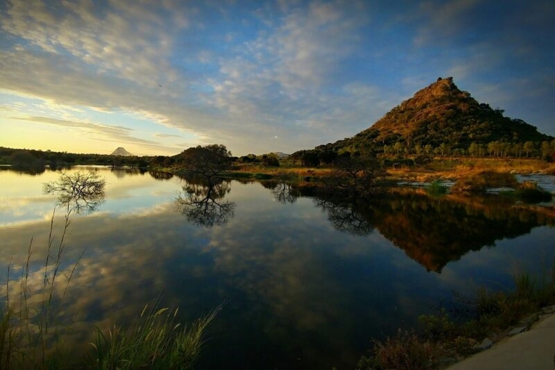 A serene lake reflects a pyramid-shaped hill and clouds during a golden hour sunset in a natural park.