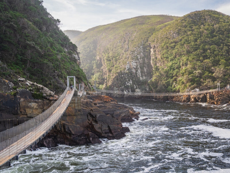 A narrow suspension bridge stretches across a rocky gorge over churning water between steep, forested hills.