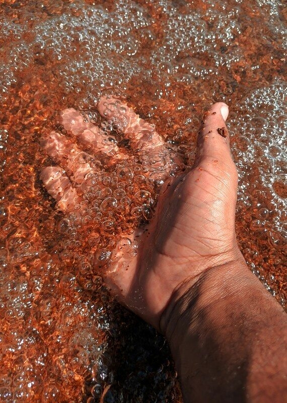 A person's hand underwater in a shallow area filled with white bubbles over a textured red-stone ground.