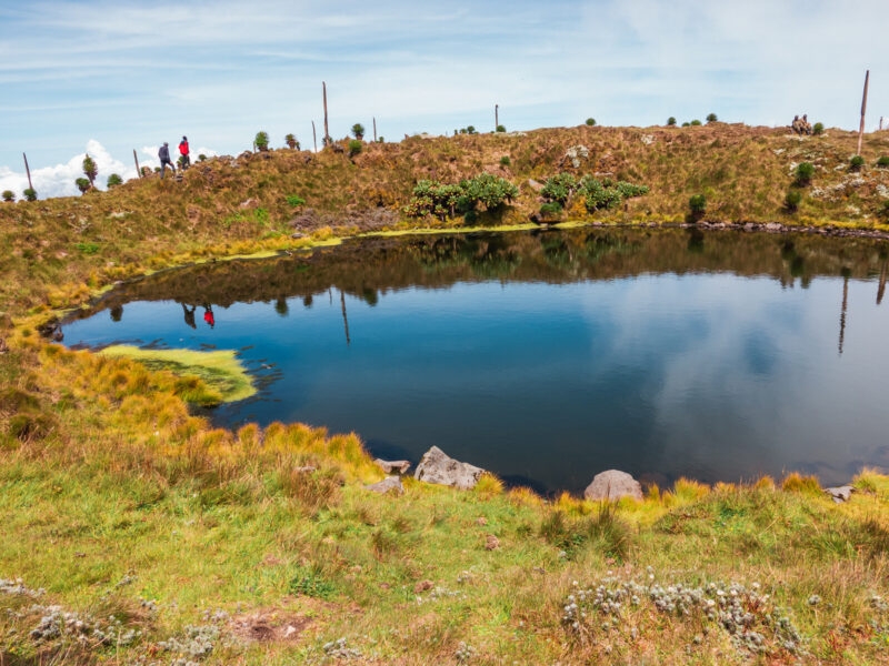 A still crater lake reflecting the blue sky, surrounded by grassy banks and hikers on the ridge.