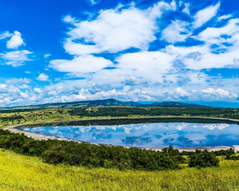 Wide view of a calm crater lake reflecting a cloudy blue sky, framed by green grassy ridges.